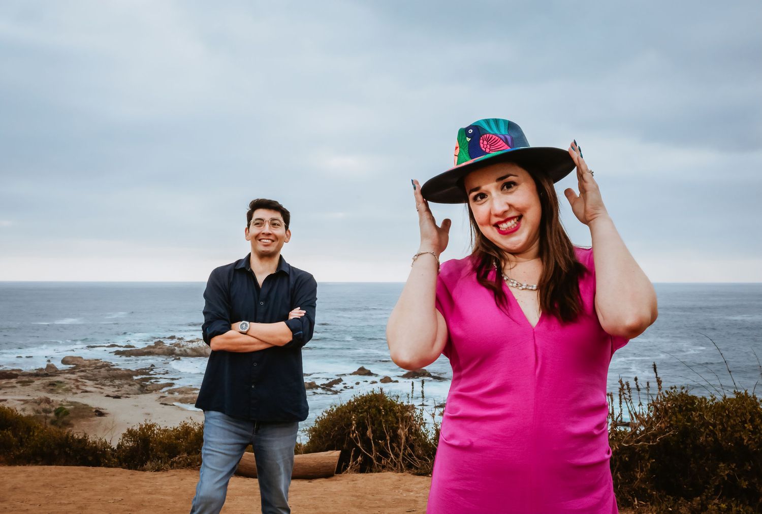 Couple posing outdoors near ocean cliffs, woman in pink dress and decorative hat, man in navy shirt smiling behind her.