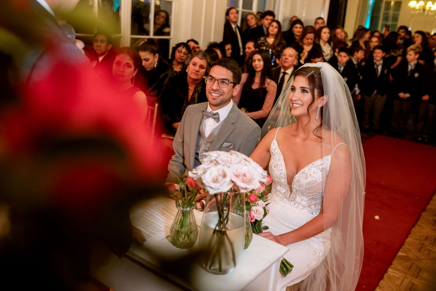Bride and groom smiling at wedding ceremony altar, bride holding pink bouquet, guests seated in elegant venue.