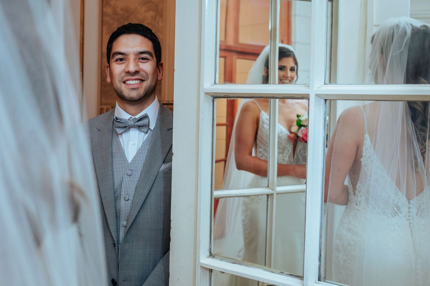 Groom in gray suit smiling at door while bride in white wedding dress holds bouquet reflected in window.