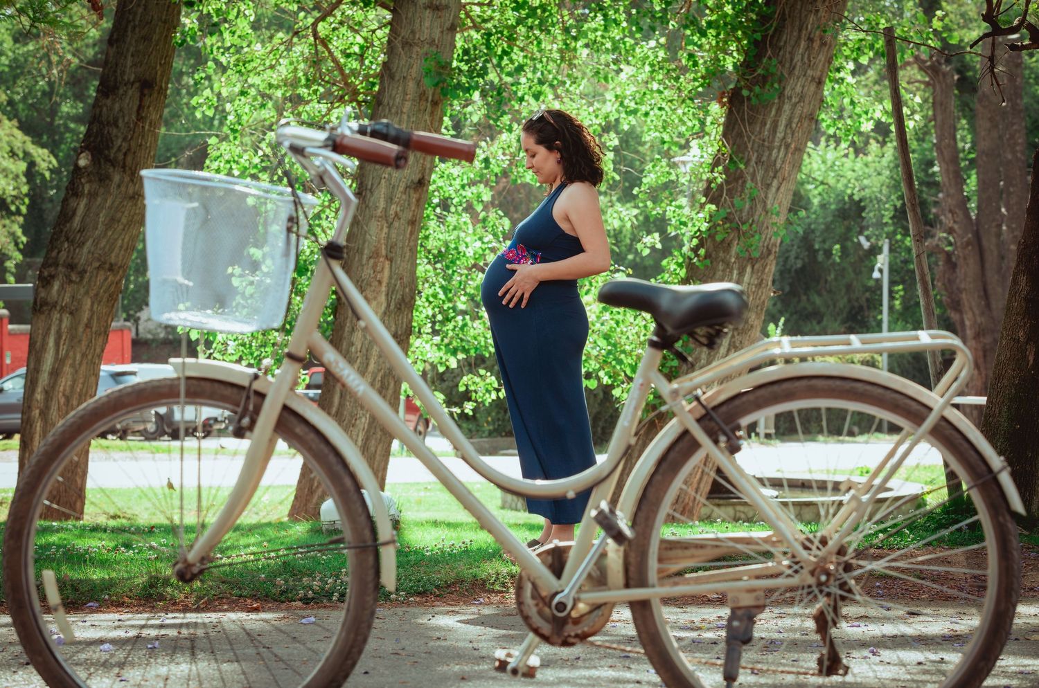 Pregnant woman in blue dress standing beside a vintage bicycle with basket in a tree-lined park.