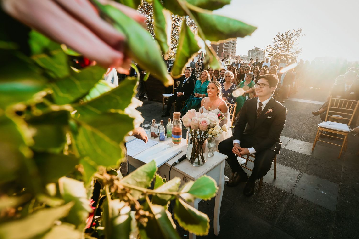 Bride and groom seated at outdoor wedding ceremony captured through green foliage.