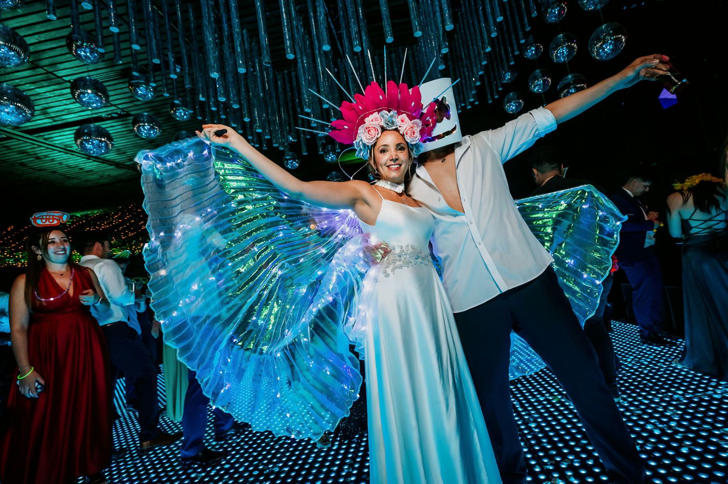 Bride wearing colorful LED wings and pink feather headpiece dances with groom at vibrant wedding reception.