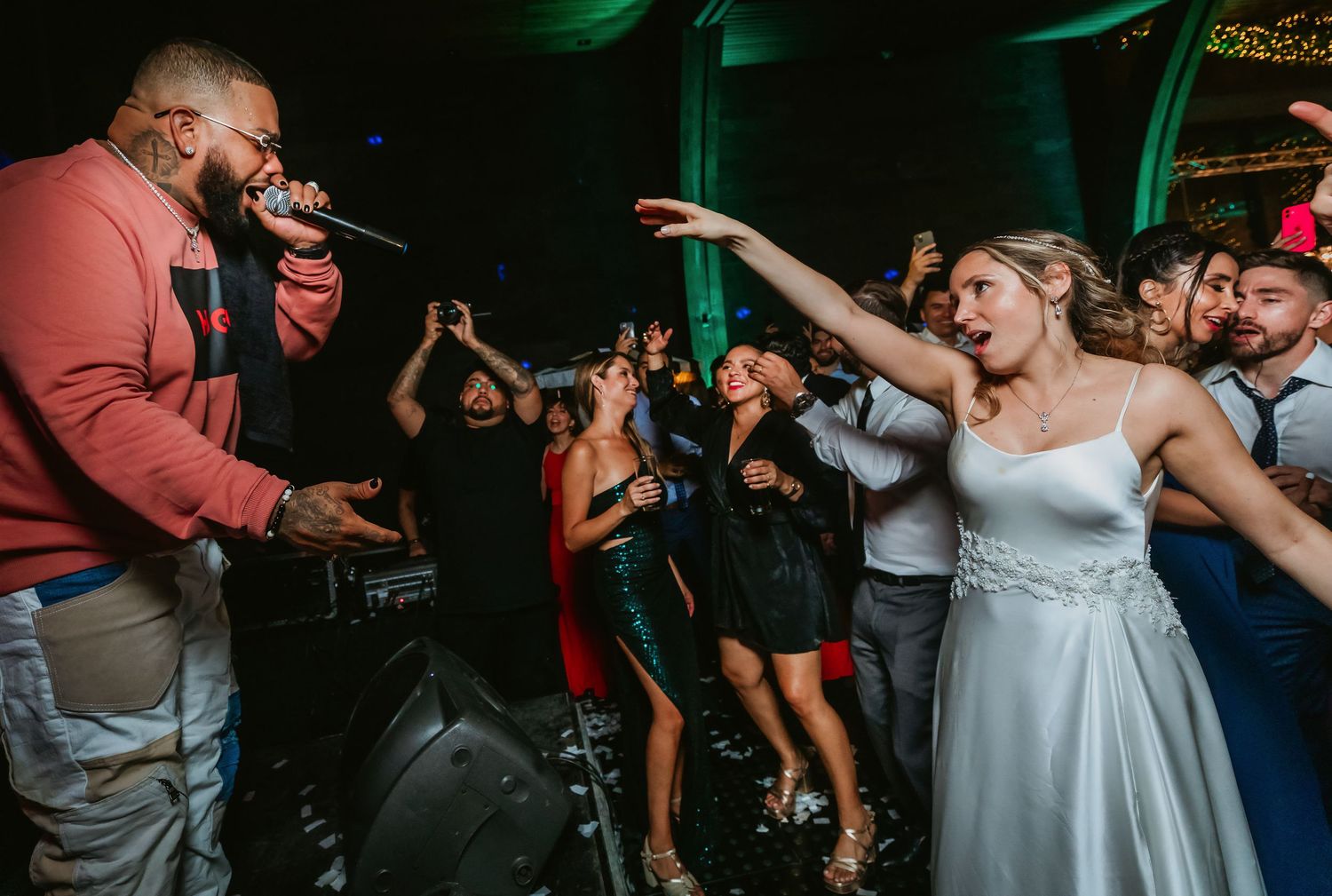 Bride in white gown dancing with performer holding microphone at lively wedding reception party.