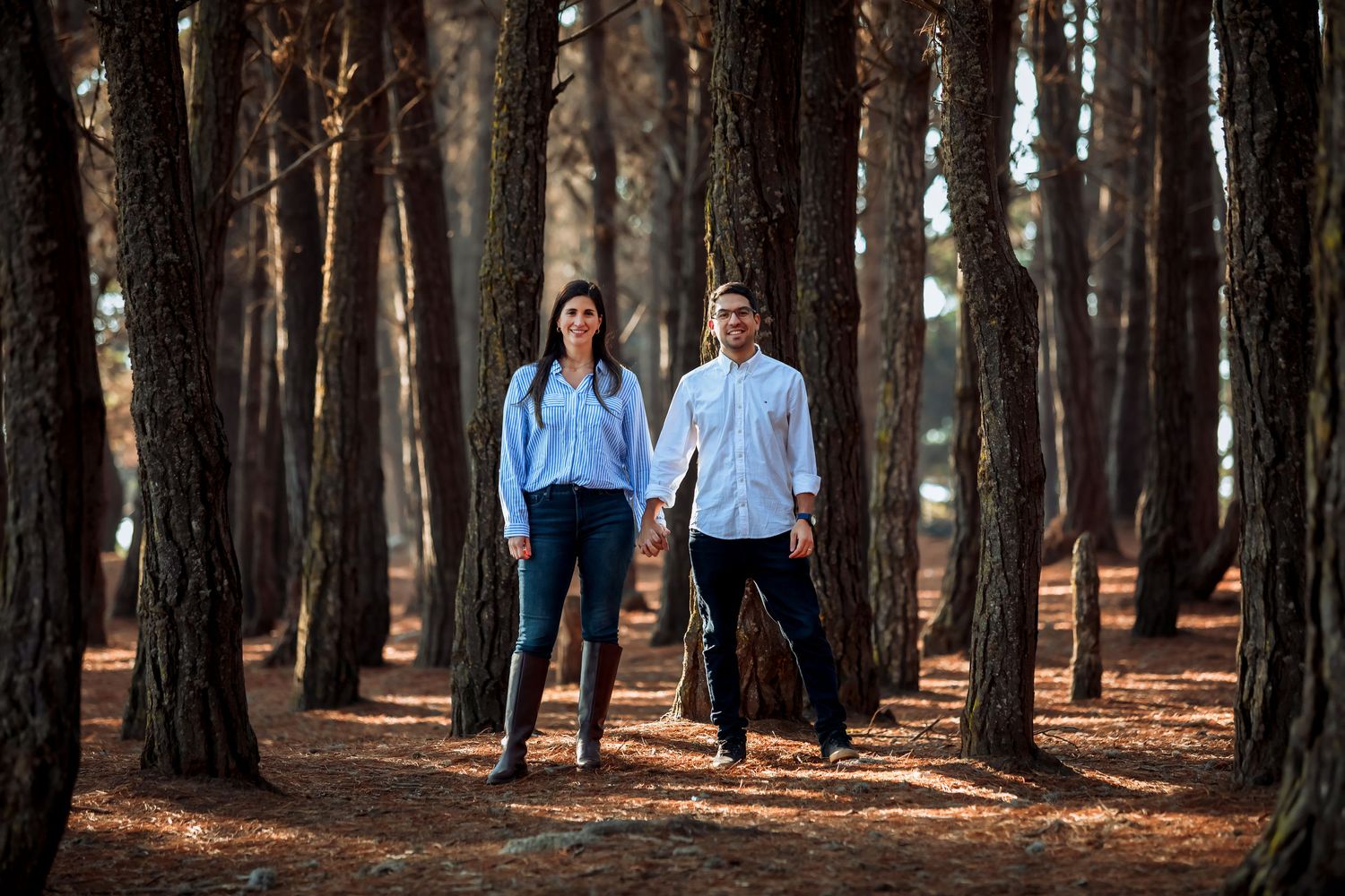 A couple holding hands and walking through a tall pine forest with a red needle-covered ground.