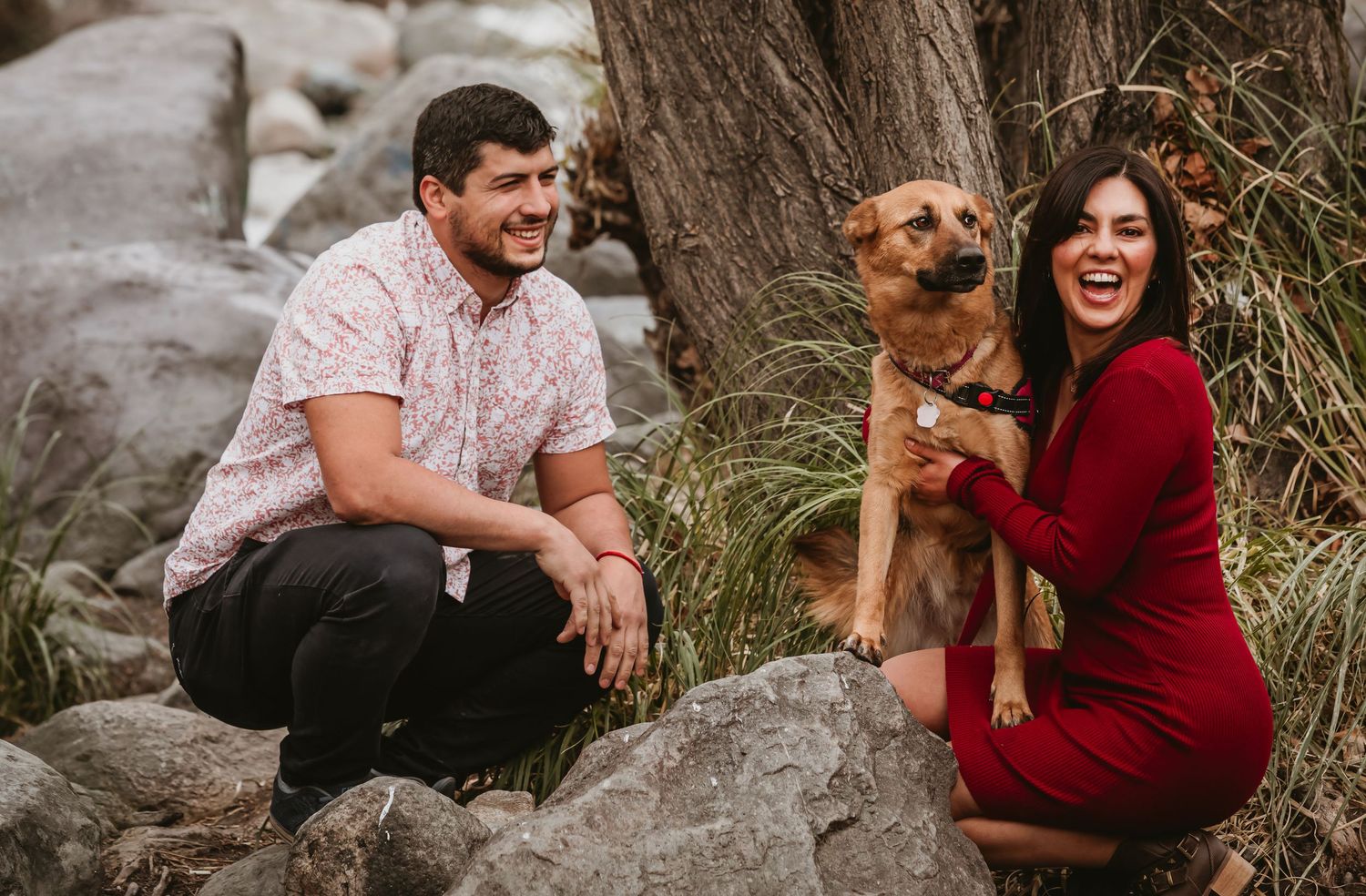Happy couple crouching with their brown dog on rocks in a natural outdoor setting.