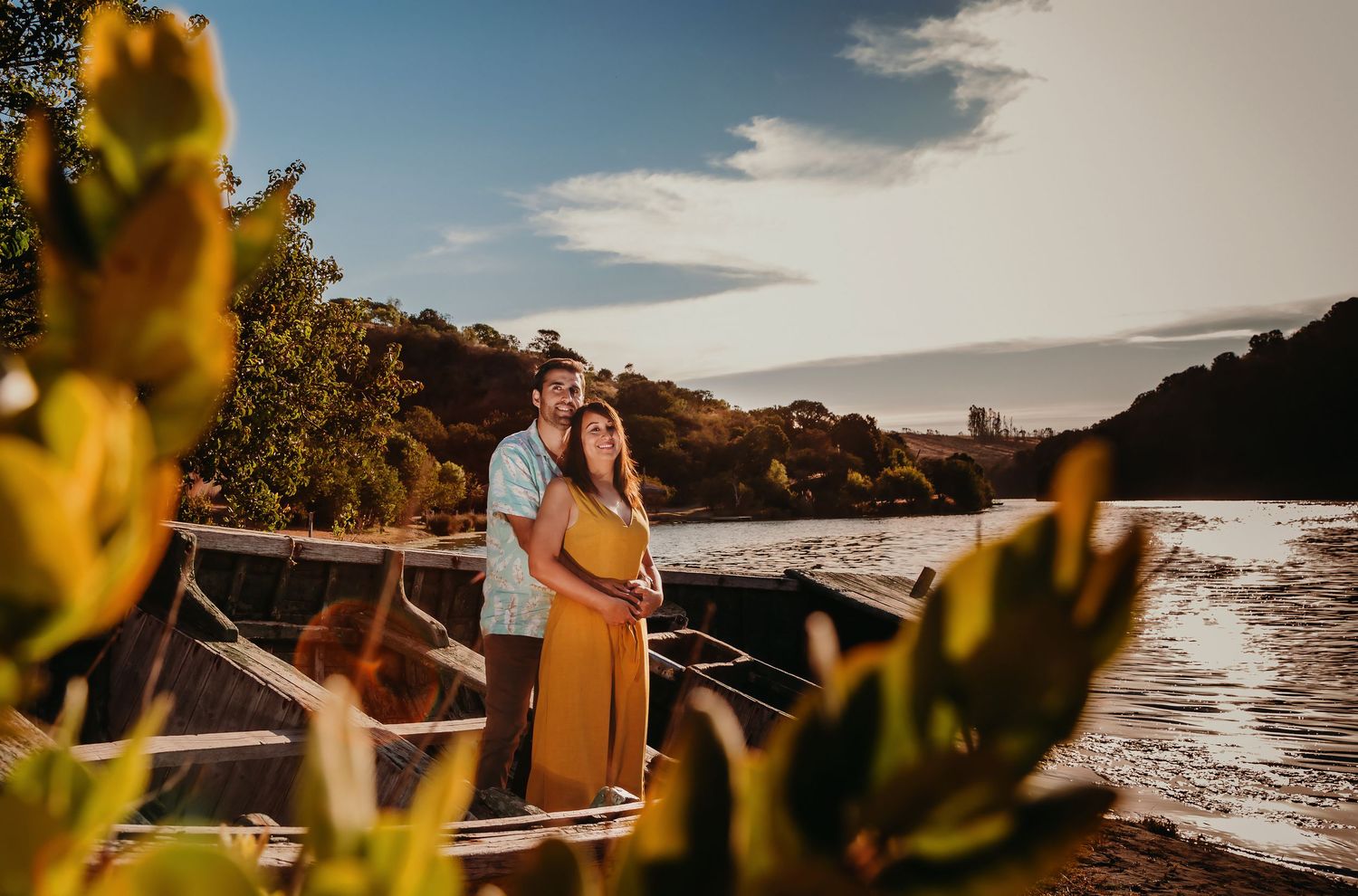 Couple embracing near an old wooden boat by a river at golden hour sunset.