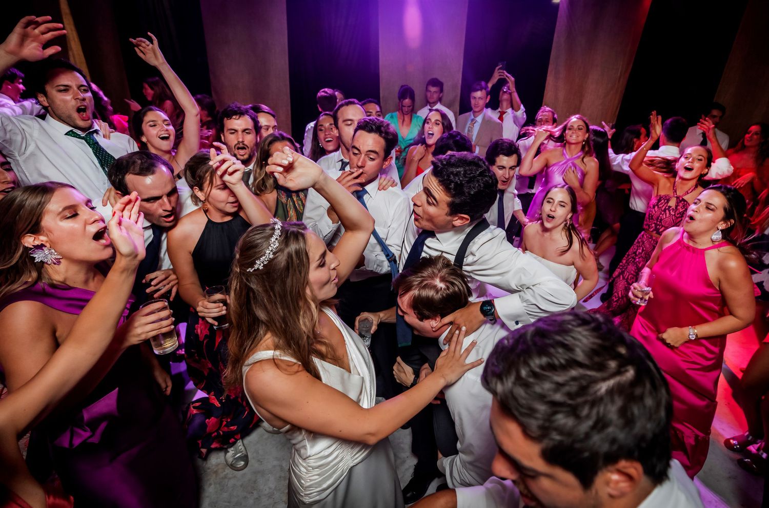 Energetic wedding reception dance floor packed with guests celebrating in colorful formal attire under pink lights.