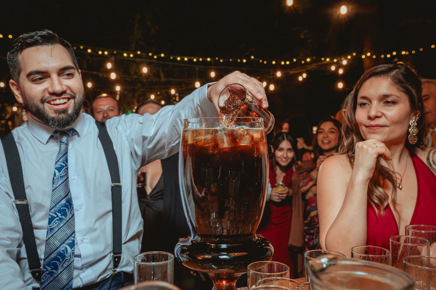 Groom pours drink into large glass dispenser at outdoor wedding reception with string lights.