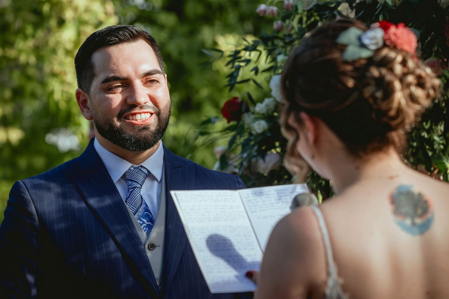 Groom smiles warmly at bride during outdoor wedding ceremony with lush green trees in background.