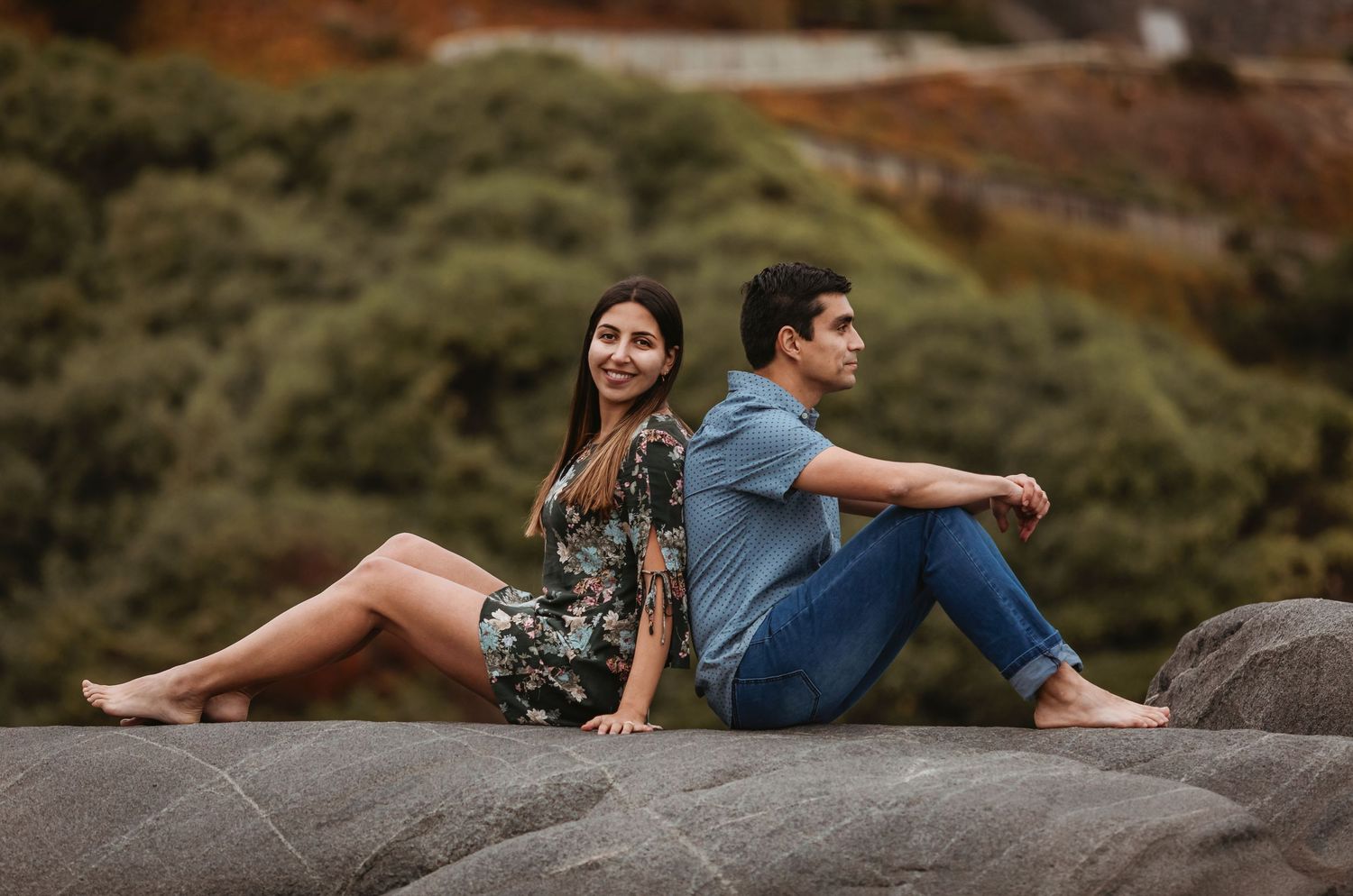 Couple sitting back to back on smooth rocks with green hillside foliage behind them.
