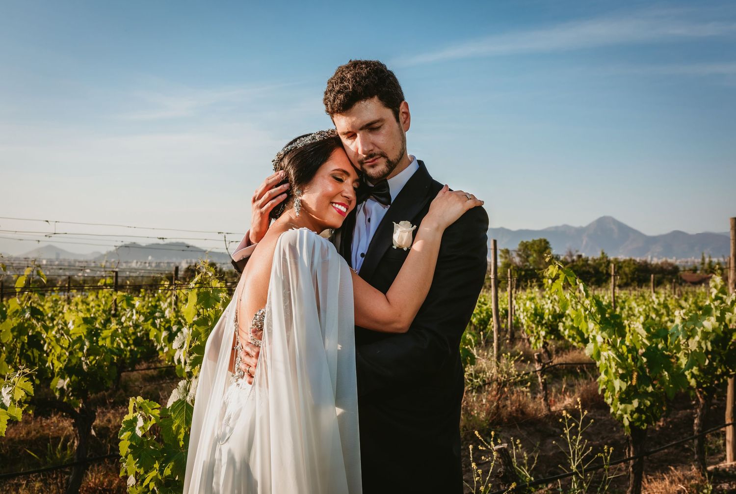 Bride and groom embracing tenderly in a lush vineyard with mountains in the background on their wedding day.