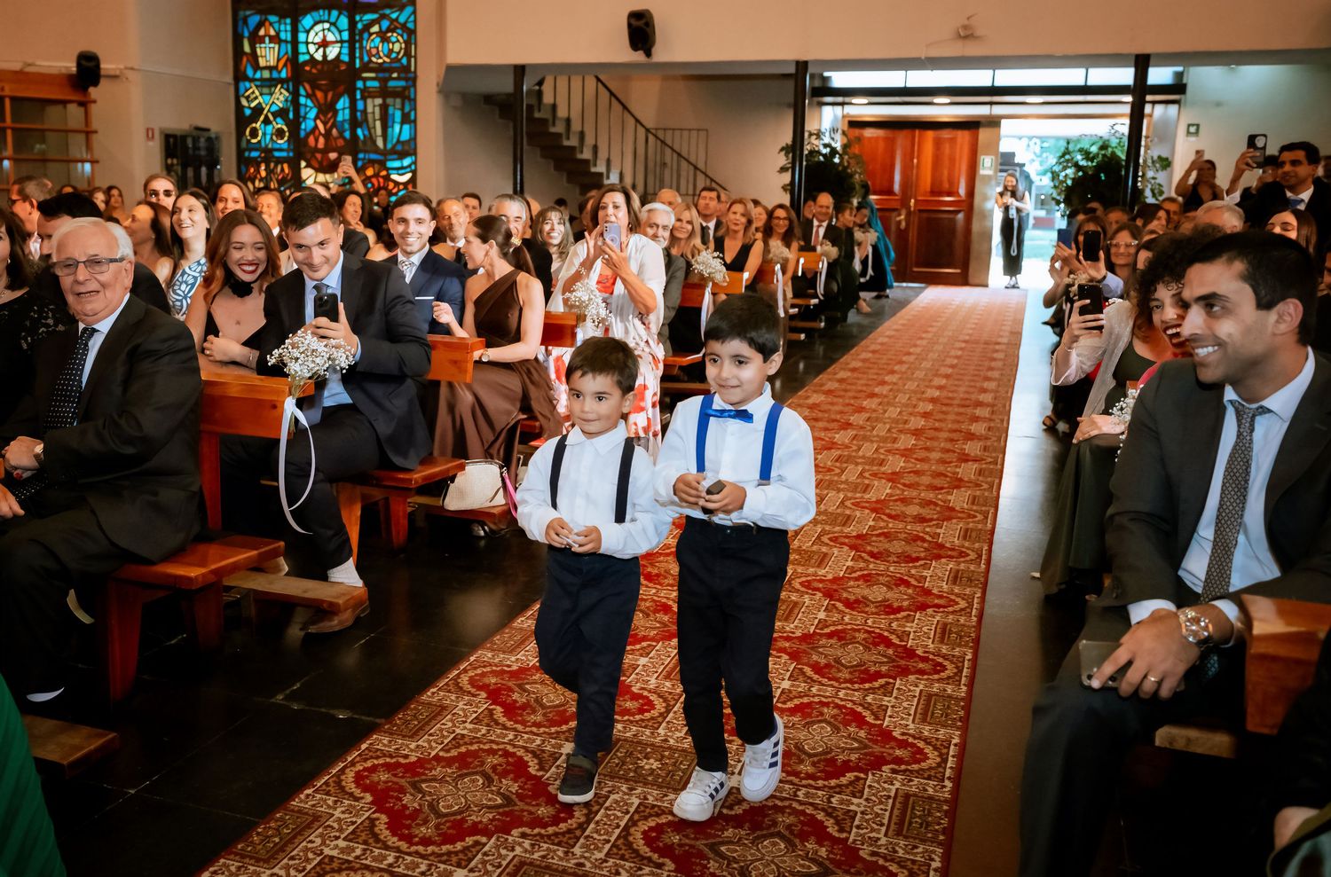 Two young ring bearers walking down church aisle during wedding ceremony while guests watch and smile.