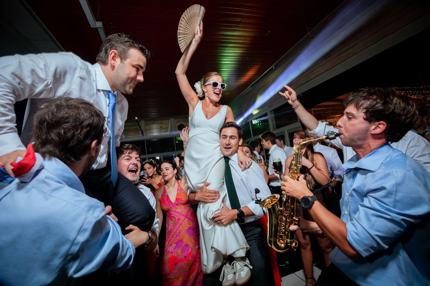 Bride lifted by guests at lively wedding reception with saxophonist and dancing crowd.