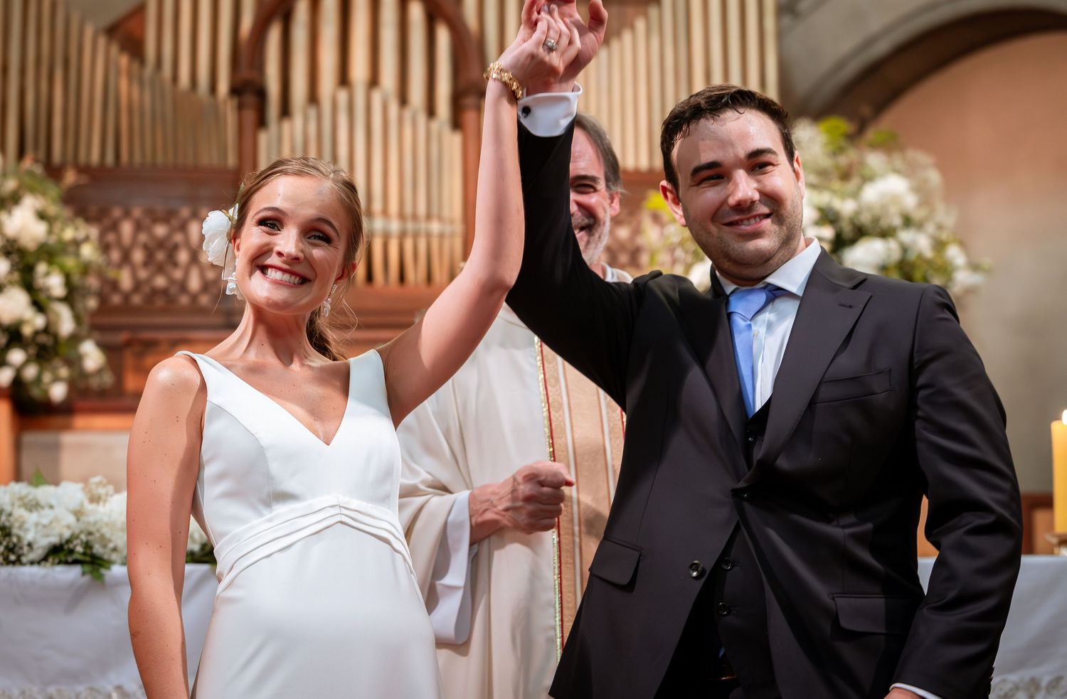 Priest raises hands of smiling newlyweds in celebration at church altar after wedding.
