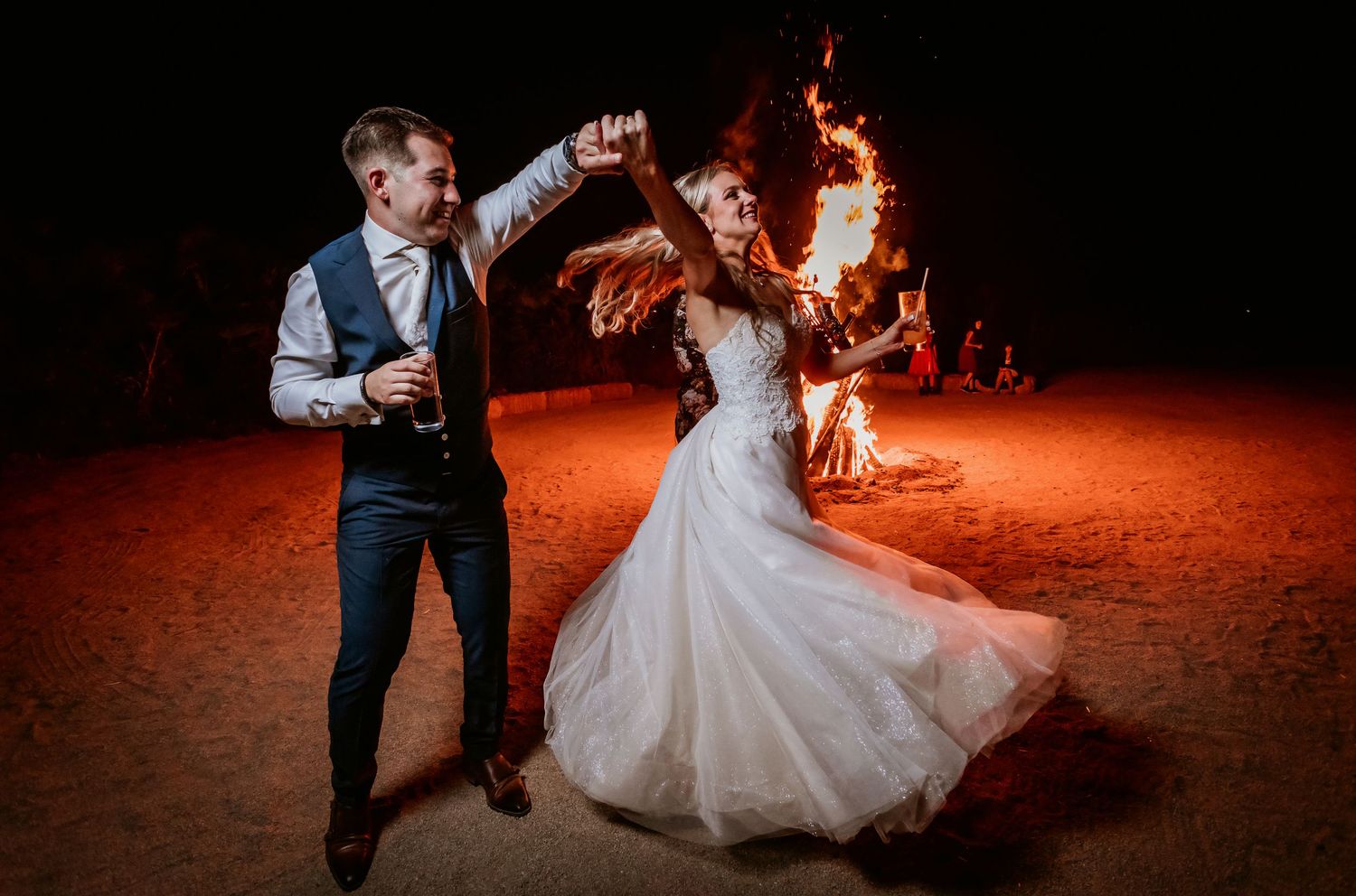 Bride and groom dancing joyfully beside a roaring bonfire during their outdoor wedding celebration at night.