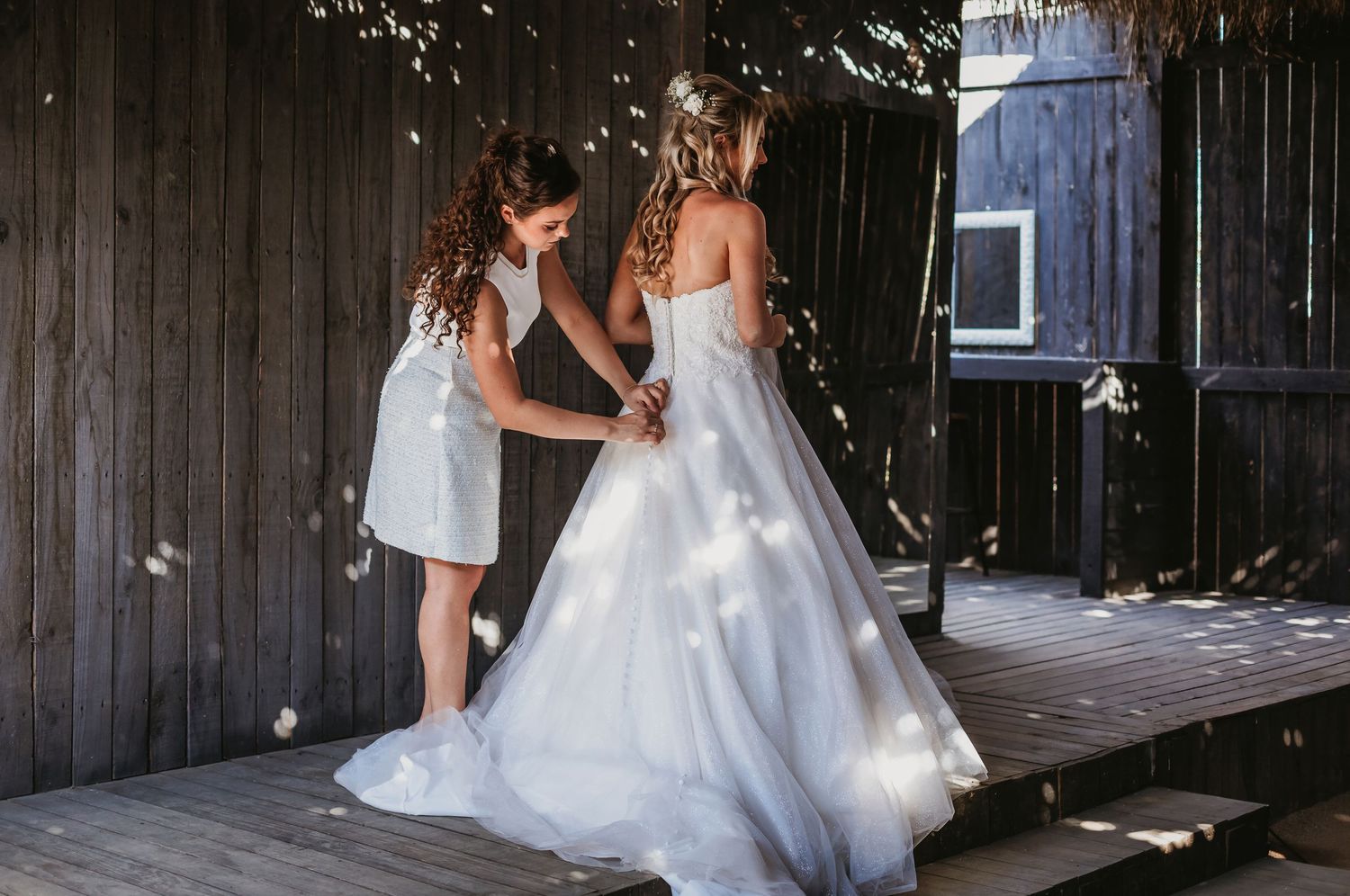 Young girl helping bride with white wedding gown on rustic wooden deck outdoors.
