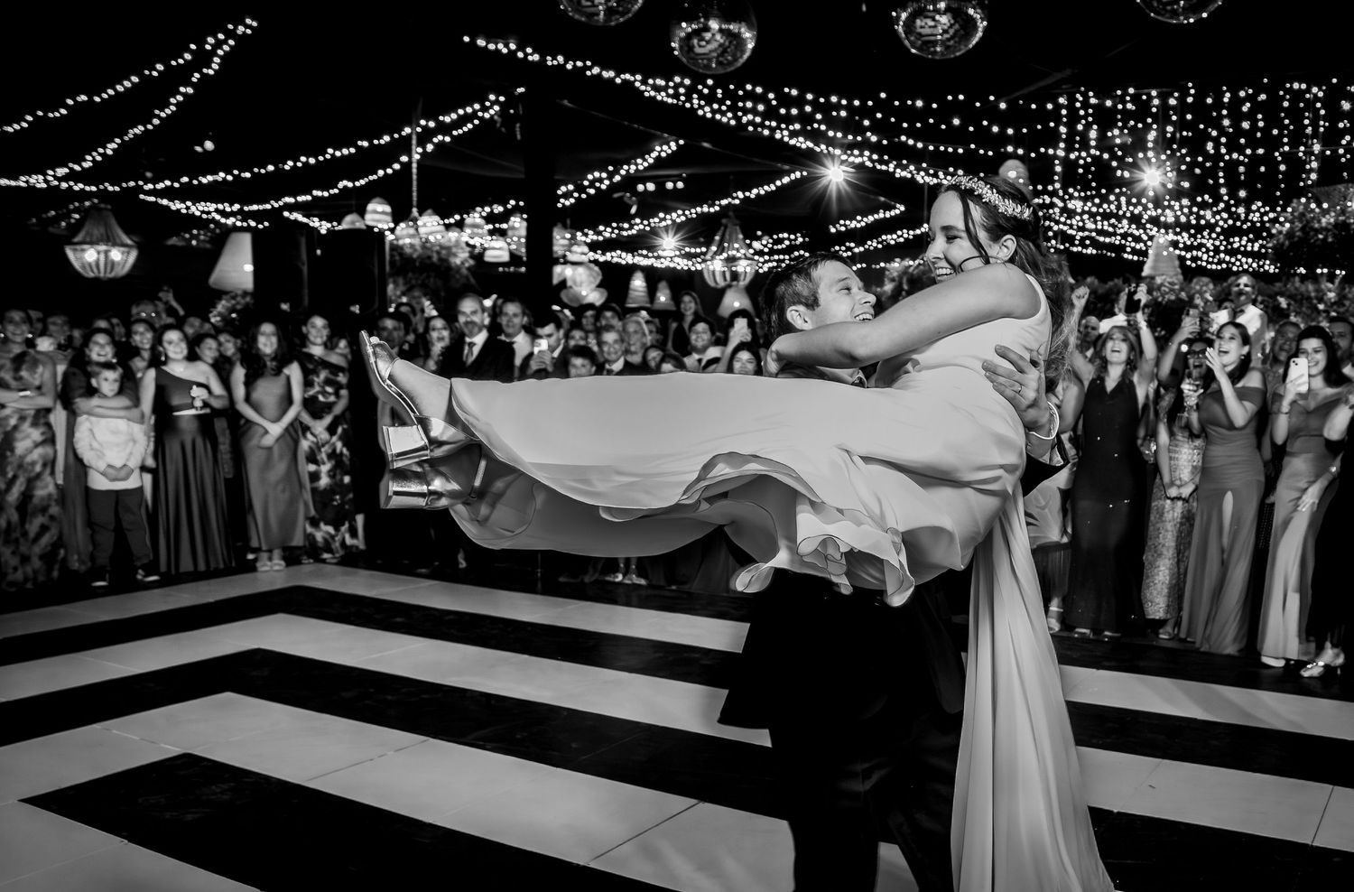 Black and white photo of groom lifting bride during first dance at wedding reception with guests watching.