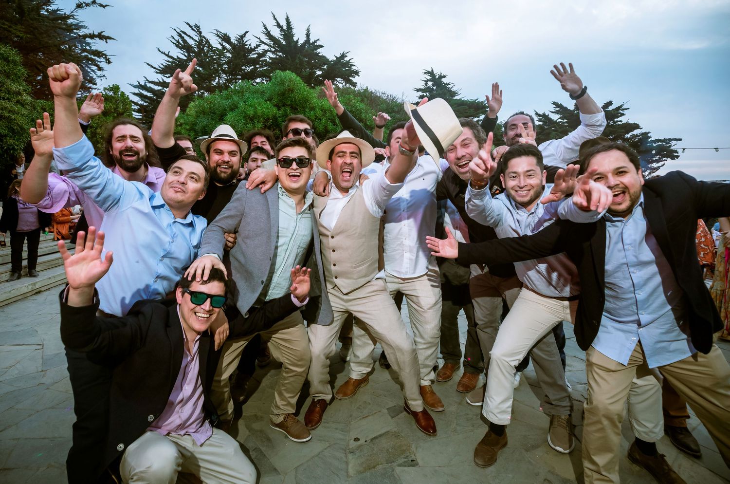 Groom celebrates joyfully with group of cheering groomsmen at outdoor wedding reception.