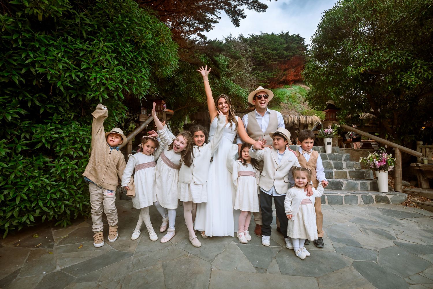 Bride and groom posing with a group of children in white outfits at an outdoor wedding.
