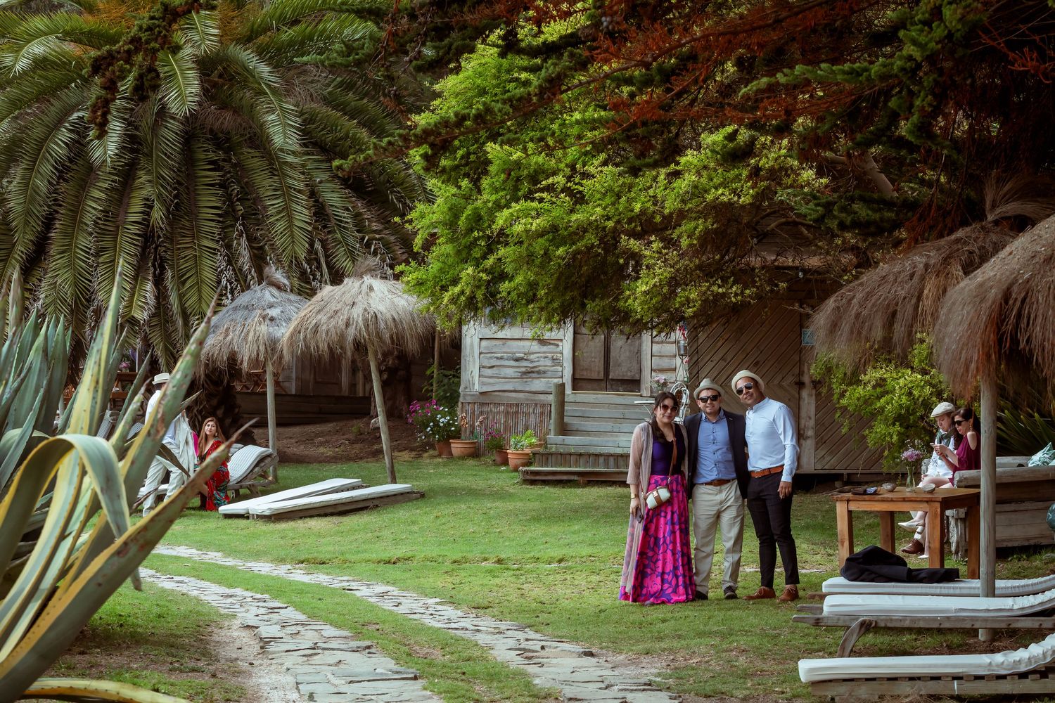 Wedding couple poses with guests near tropical thatched huts and lush greenery.