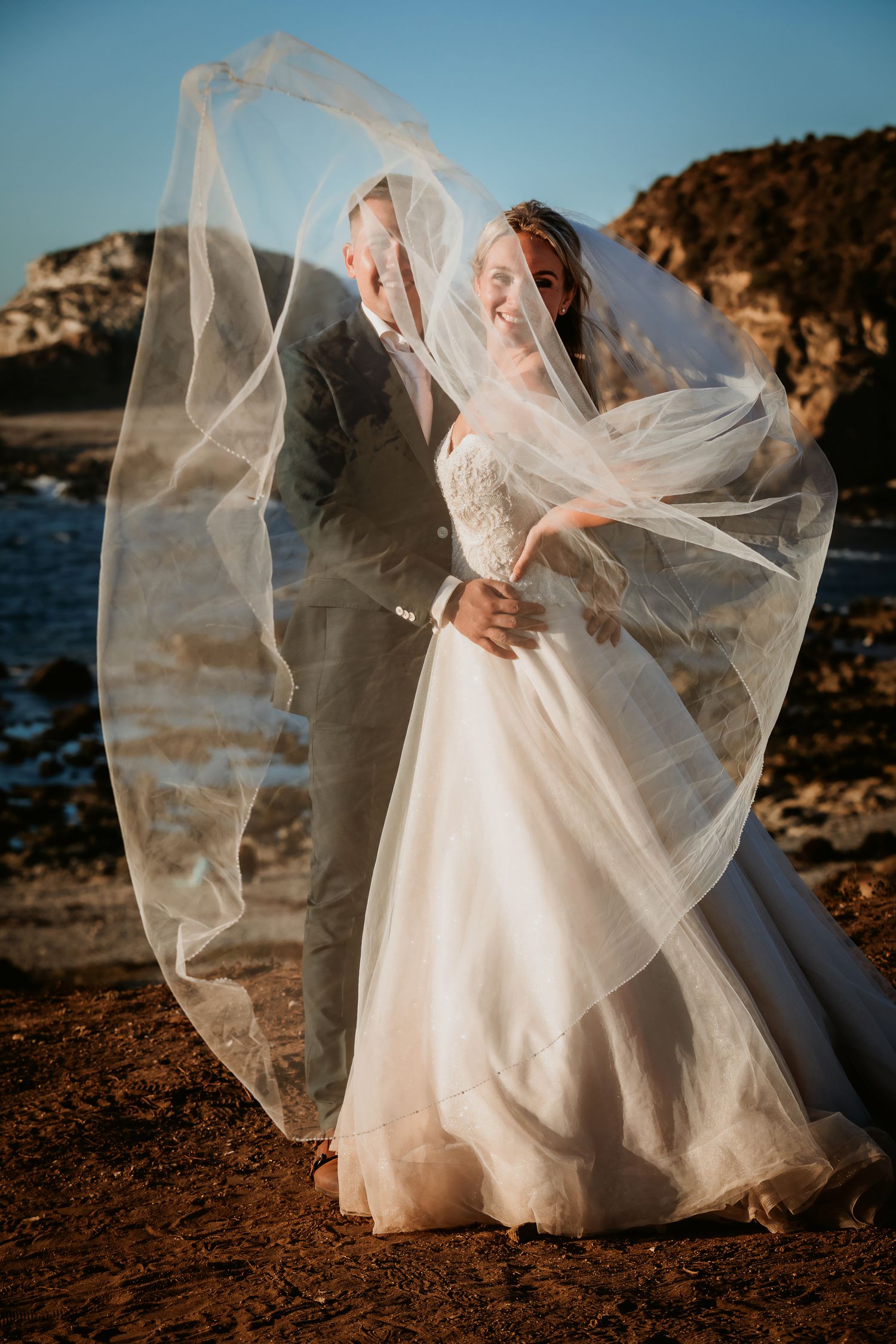 Groom partially hidden by bride's flowing veil as couple laughs together on rocky ocean shoreline.