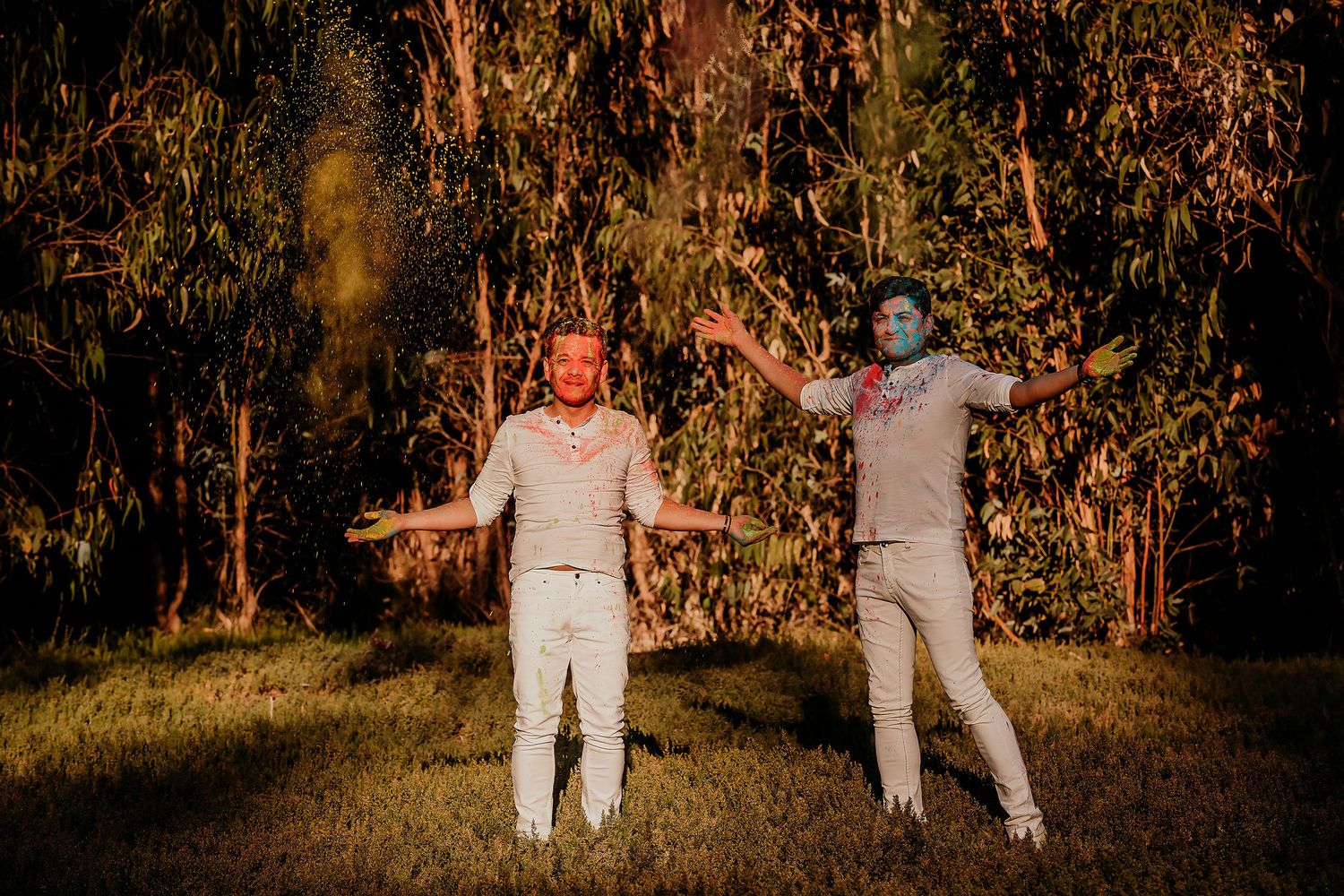 Two men covered in colorful Holi powder stand with arms outstretched in a green outdoor setting.
