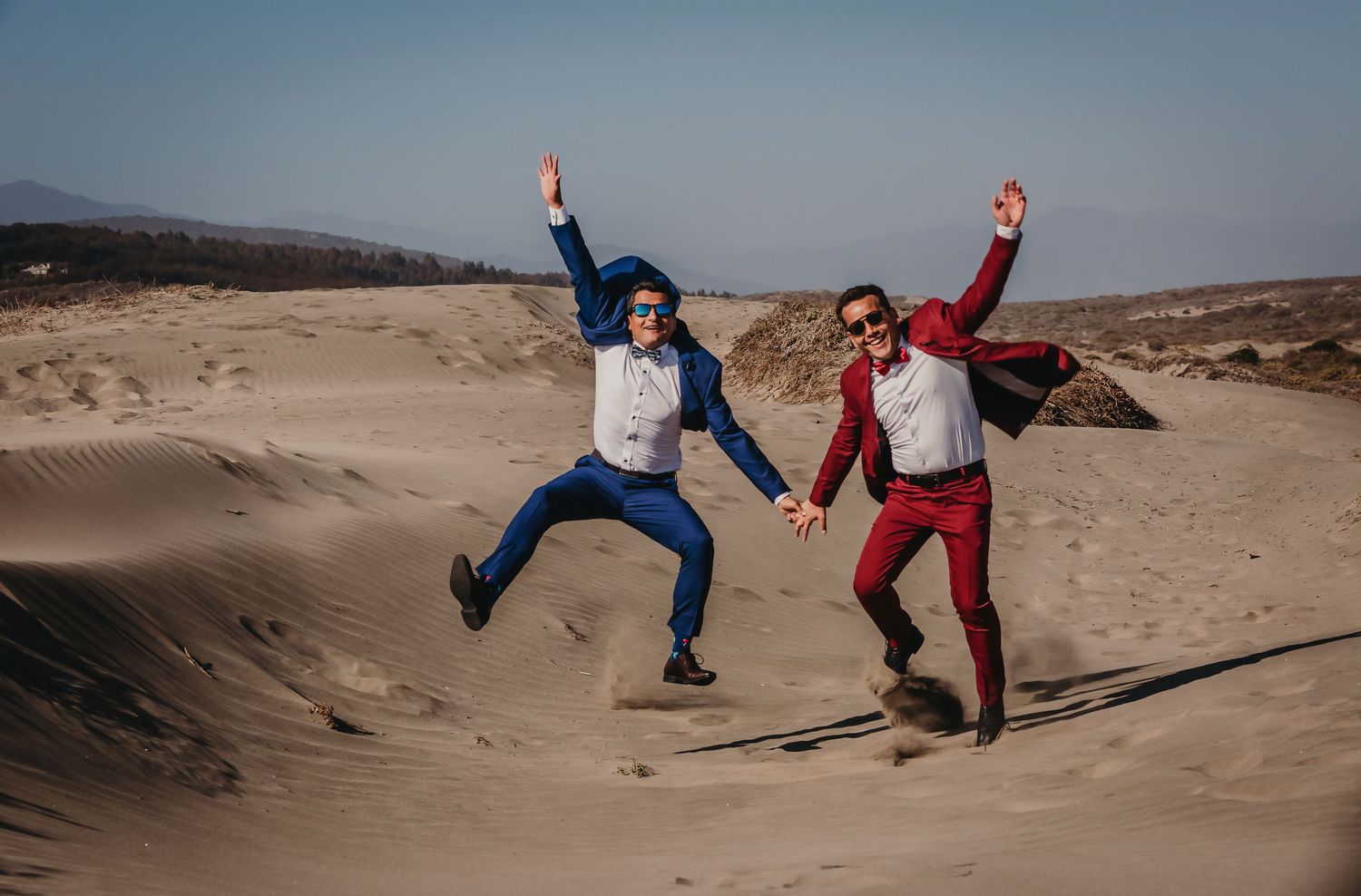 Two grooms in red and blue suits jumping joyfully on sandy desert dunes under a clear blue sky.