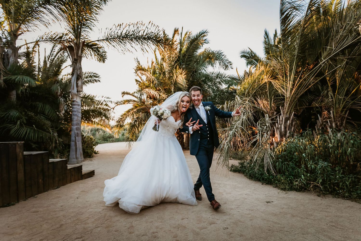 Joyful bride and groom walking on sandy beach path surrounded by tropical palm trees.
