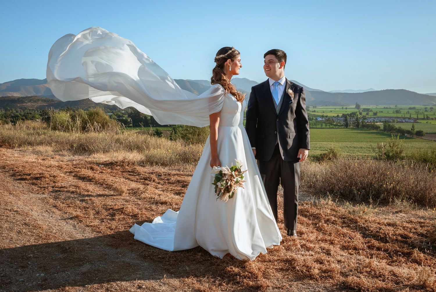 Bride with veil blowing in wind stands with groom on a rural dirt path with mountain views.