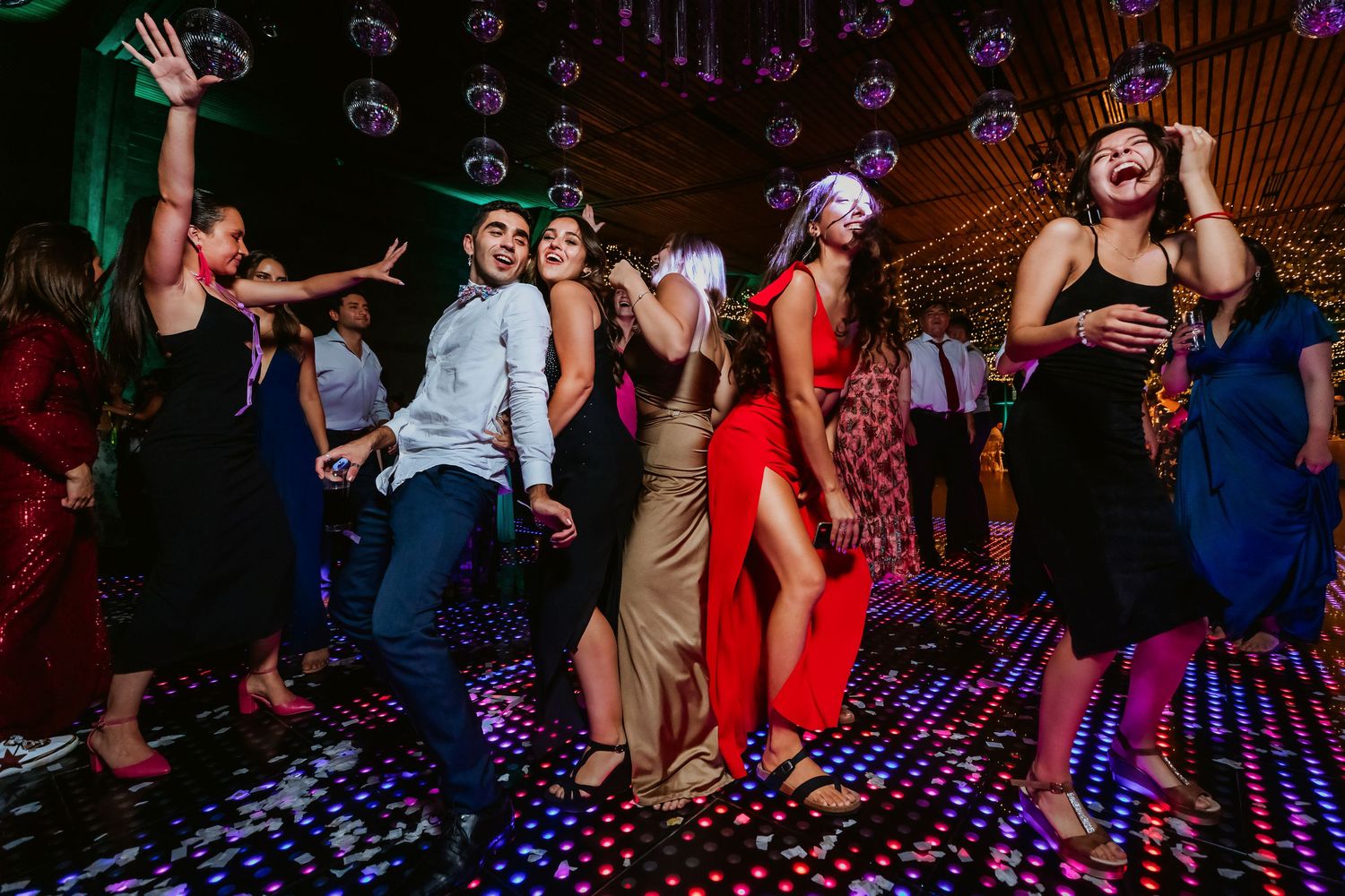 Guests in elegant attire dancing on colorful LED dance floor at wedding reception.