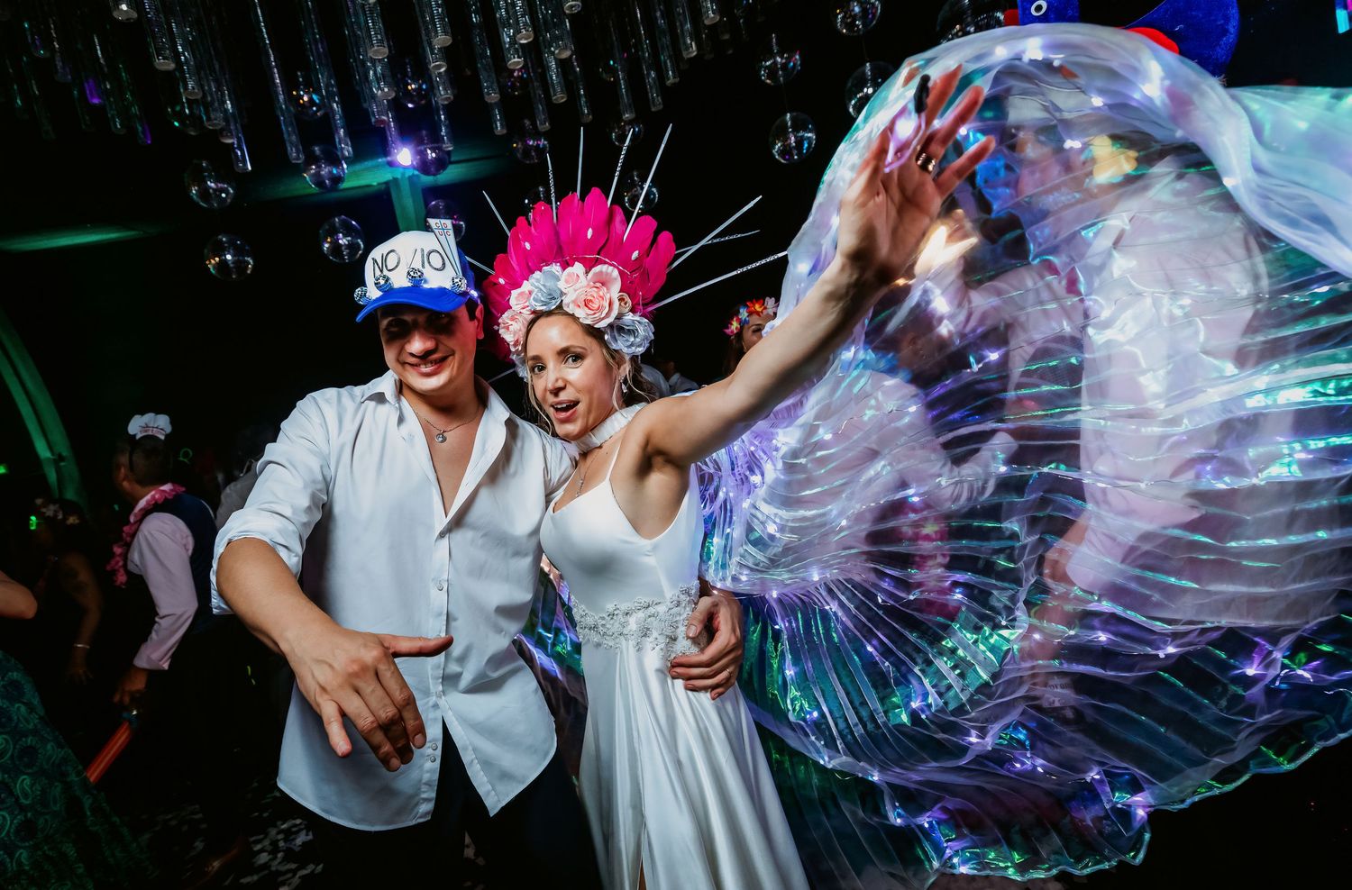 Bride and groom posing with illuminated butterfly wings and colorful headpieces at wedding reception.