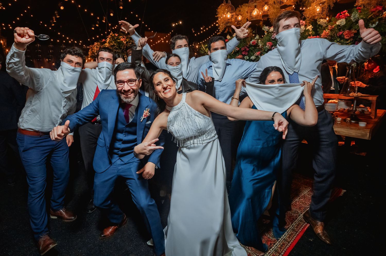 Wedding party playfully pulls masks over faces with bride and groom on dance floor.