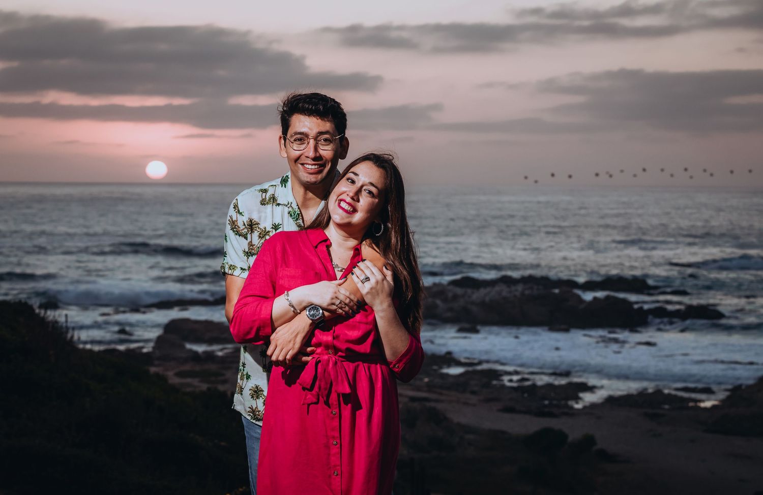 Couple embracing at sunset on rocky beach, woman in pink dress, man in floral shirt, ocean backdrop.