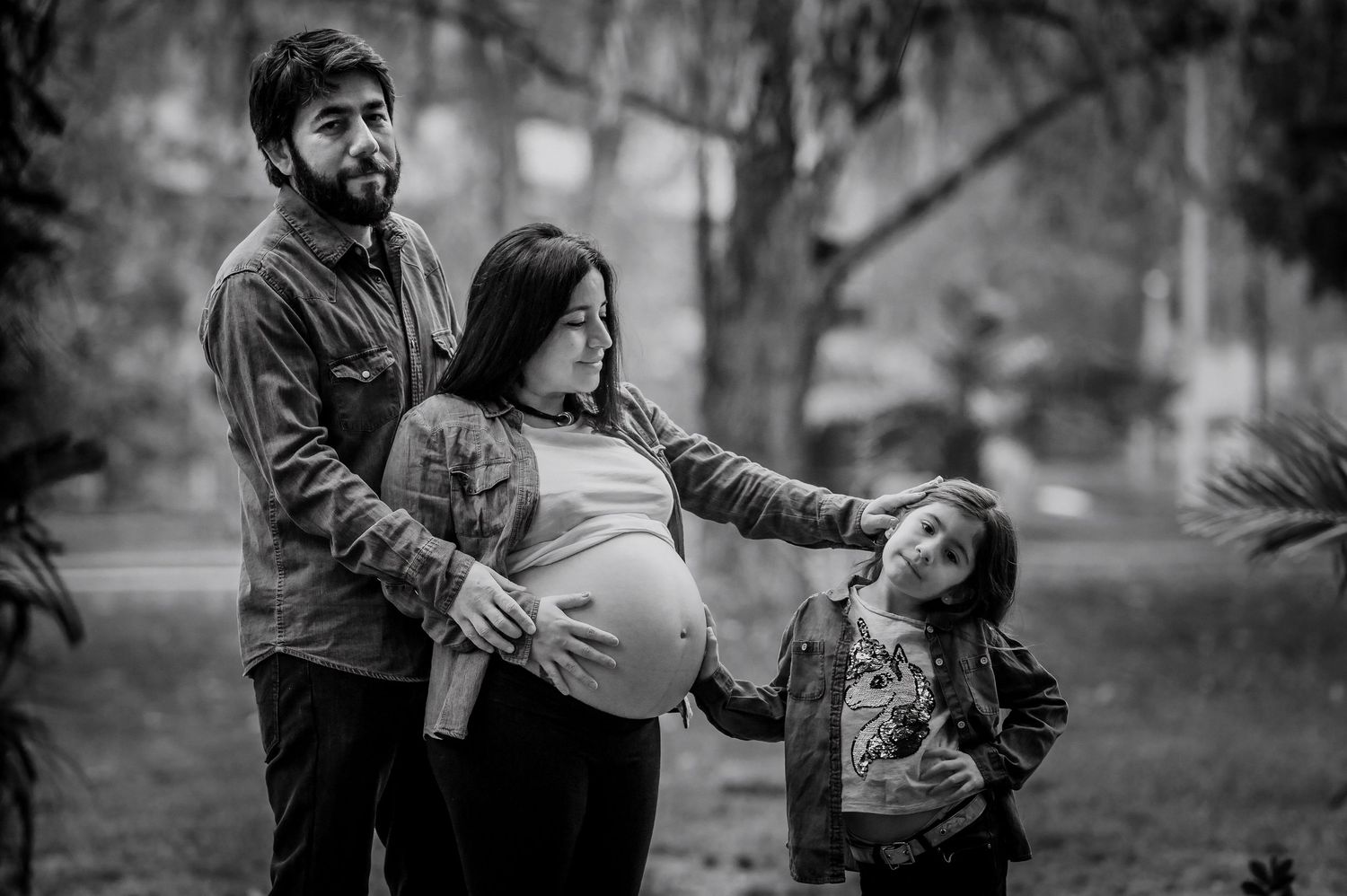 Black and white family maternity photo with father, pregnant mother, and young child in park.