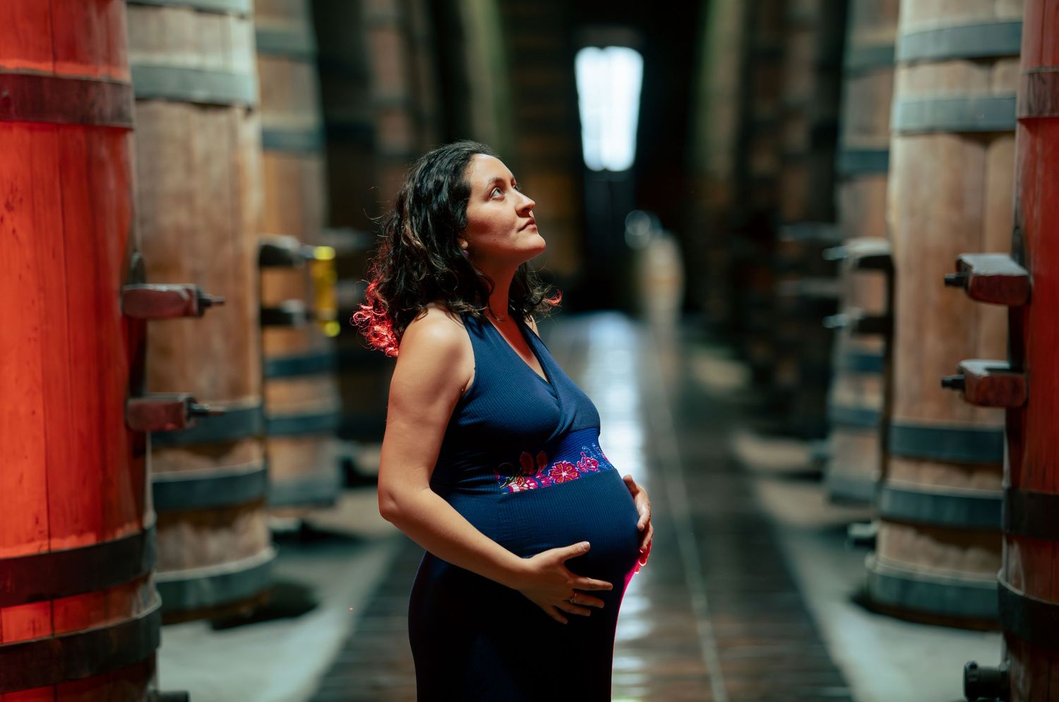 Pregnant woman in blue dress cradles baby bump among rows of wooden barrels in winery cellar.