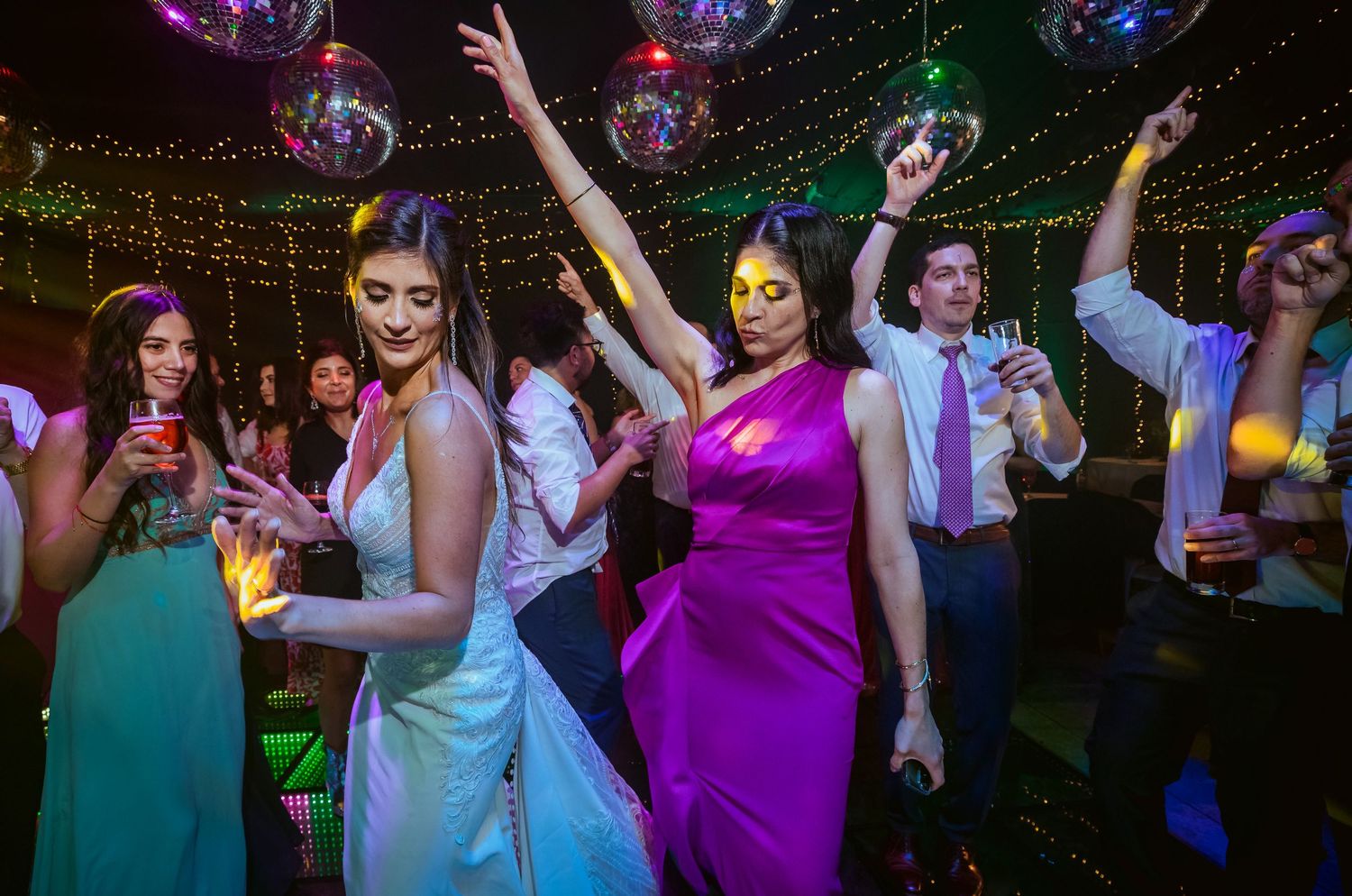 Bride and guests in colorful gowns dancing joyfully under disco balls at a lively wedding reception party.