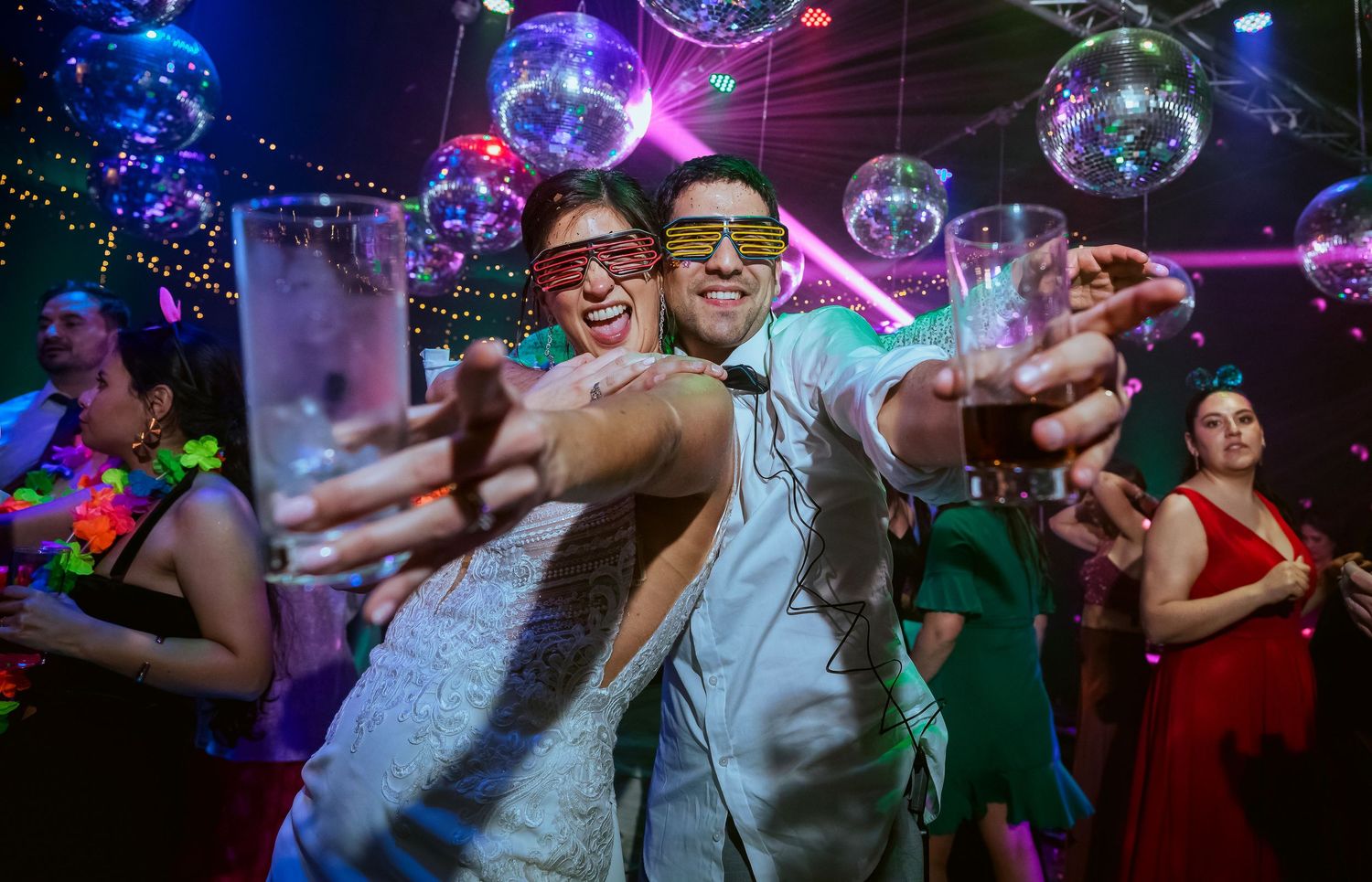 Couple in wedding attire dancing at disco party, raising glasses, with mirror ball decorations and colorful lights.