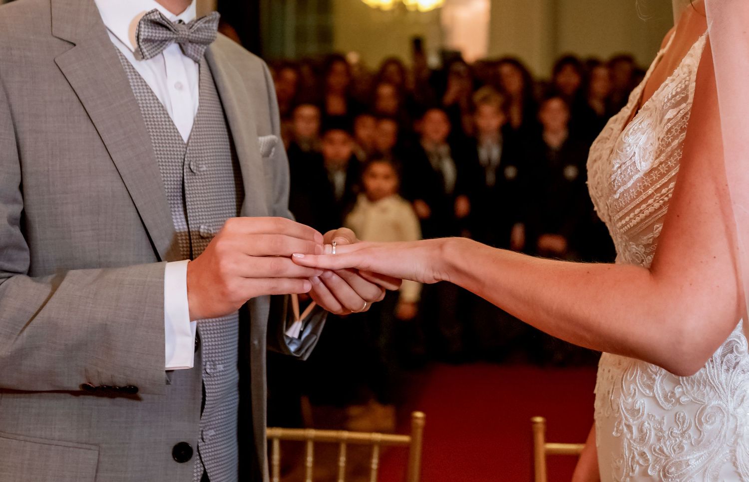 Groom placing a wedding ring on bride's finger during a romantic ceremony with guests seated in the background.