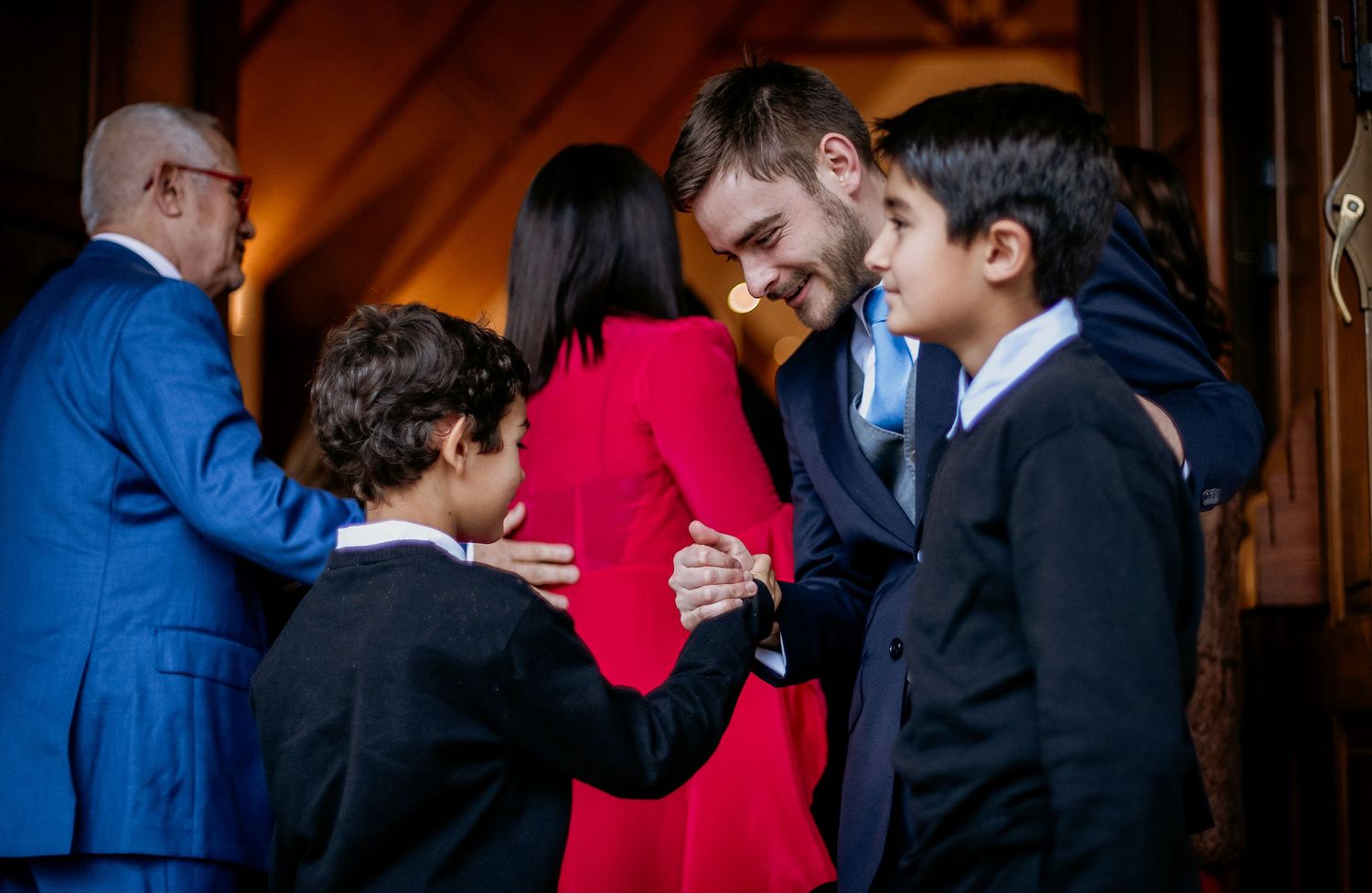 A man in a suit greets two young boys while a woman in red looks on at a formal gathering.