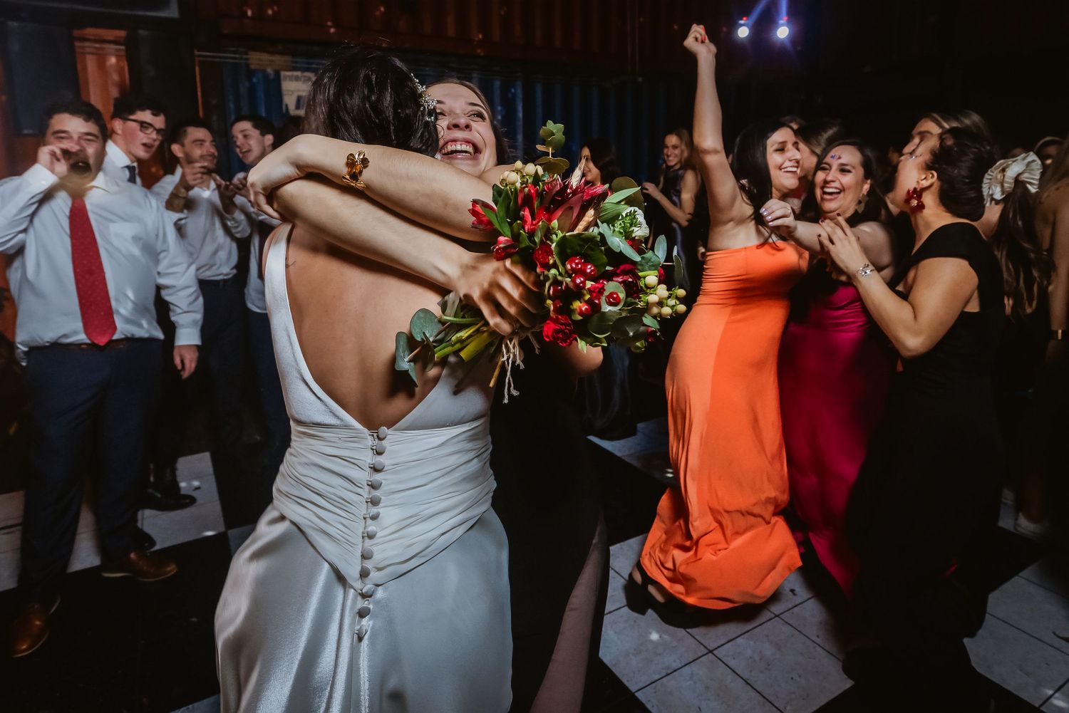 Bride hugging a guest on the dance floor while bridesmaids in orange dresses celebrate joyfully at a wedding reception.
