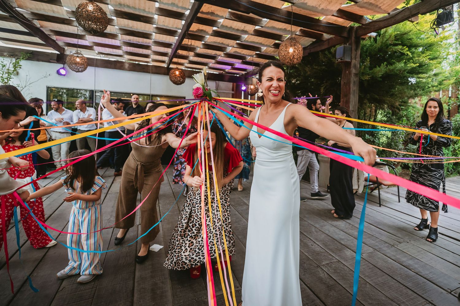 Bride in white gown laughing while holding colorful ribbons at an outdoor wedding celebration.