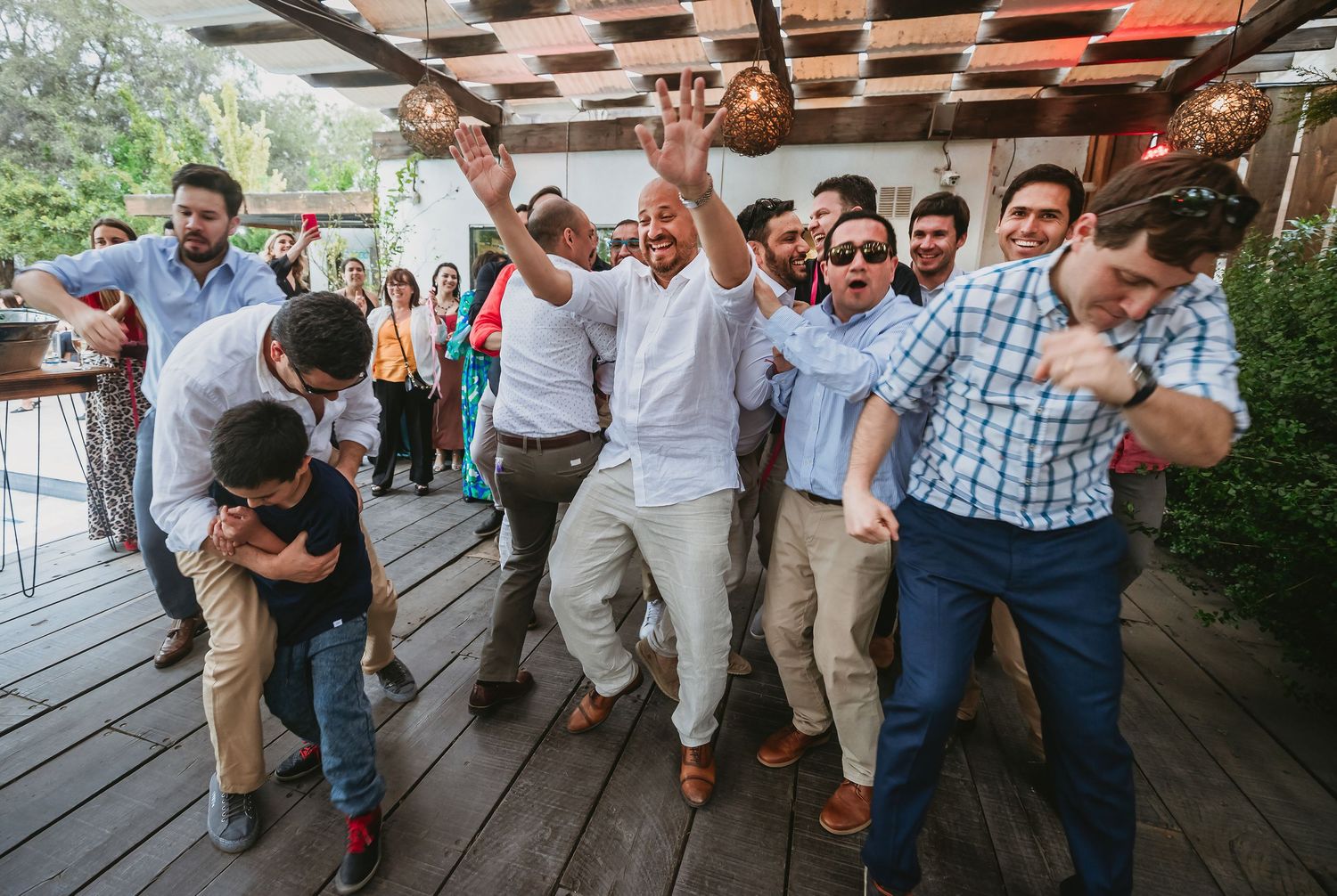Group of men dancing and celebrating outdoors on a wooden deck at a festive party or wedding reception.