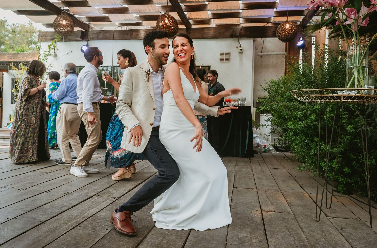 Bride and groom dancing joyfully at outdoor wedding reception on wooden deck with guests celebrating in background.