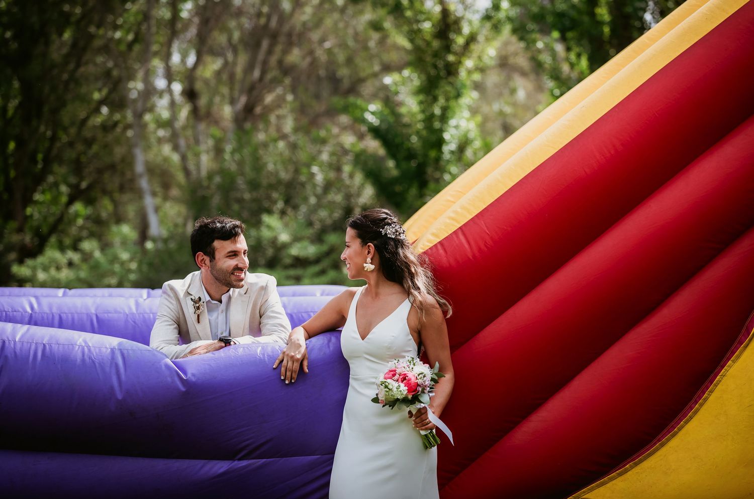 Bride and groom laughing beside a colorful inflatable bounce house outdoors at their fun wedding celebration.