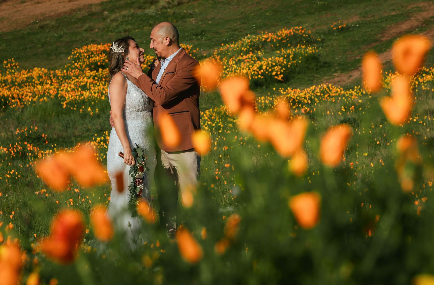 Couple embracing and smiling at each other in a field of vibrant orange California poppies during a sunny outdoor wedding portrait.