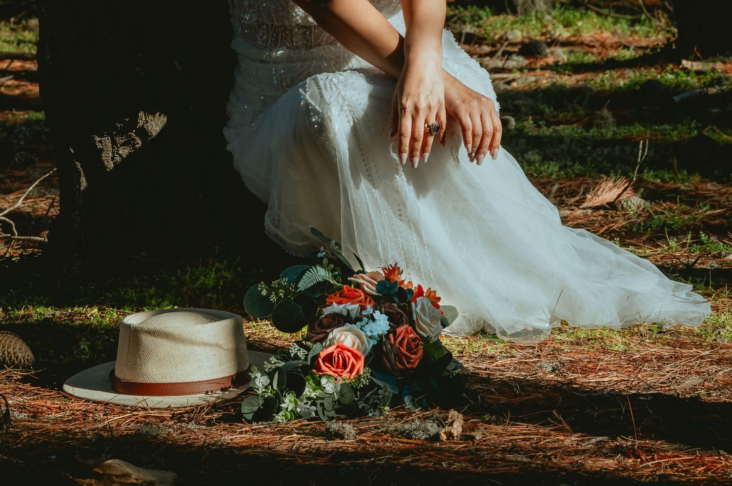 Bride in a white dress kneeling on a forest floor beside a floral bouquet and a straw hat in dappled sunlight.