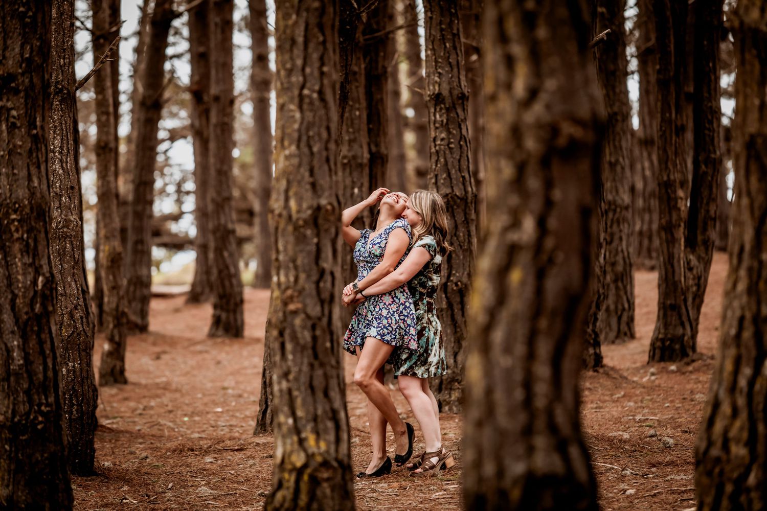 Two women laugh and dance joyfully together in a scenic pine forest setting.