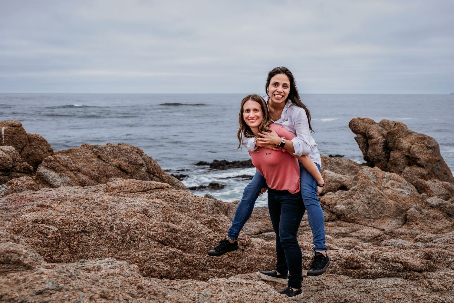 One woman gives another a piggyback ride on a rocky beach by the sea.