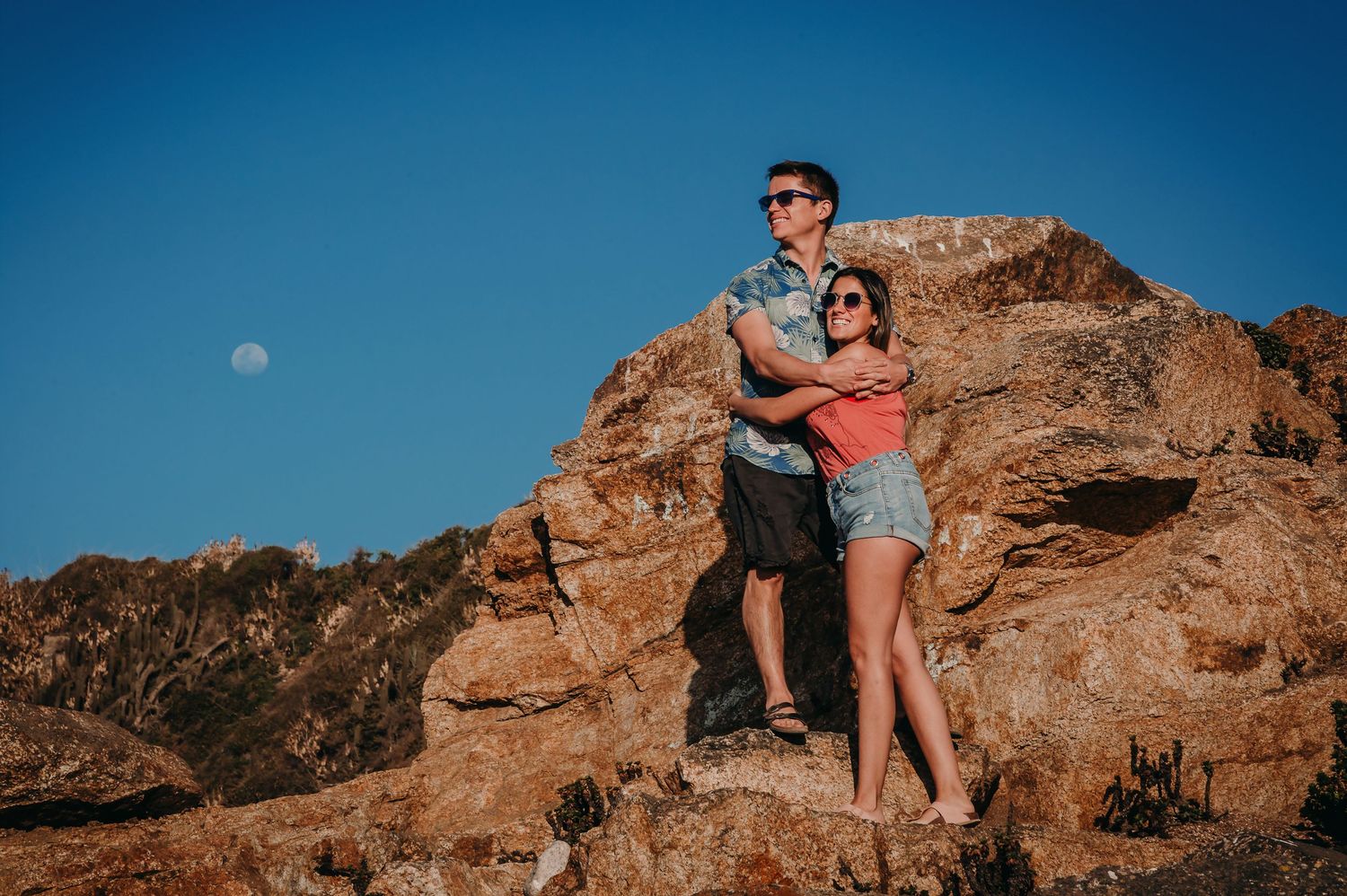Couple hugging on sun-lit rocks with moon visible in blue sky.