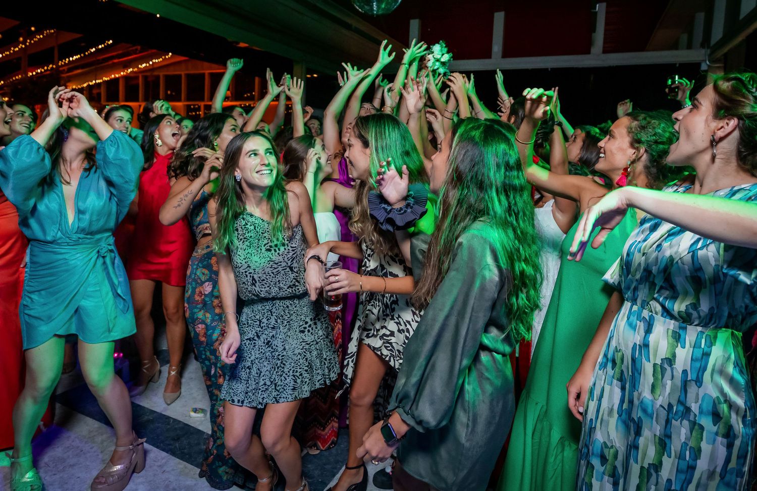 Guests in colorful dresses dance energetically under green lights at a lively wedding reception.